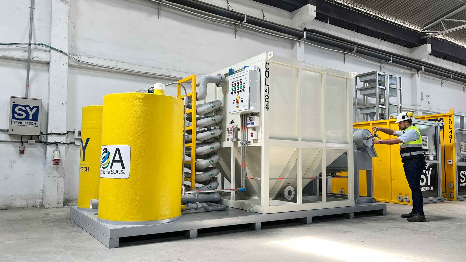Técnico inspeccionando una planta de tratamiento de aguas residuales modular de color verde marca BIOMAX SYNERTECH, que incluye módulos de pretratamiento, decantación, DAF y oxidación avanzada. La planta está ubicada en un entorno industrial con techo alto y tuberías visibles. - ver más información