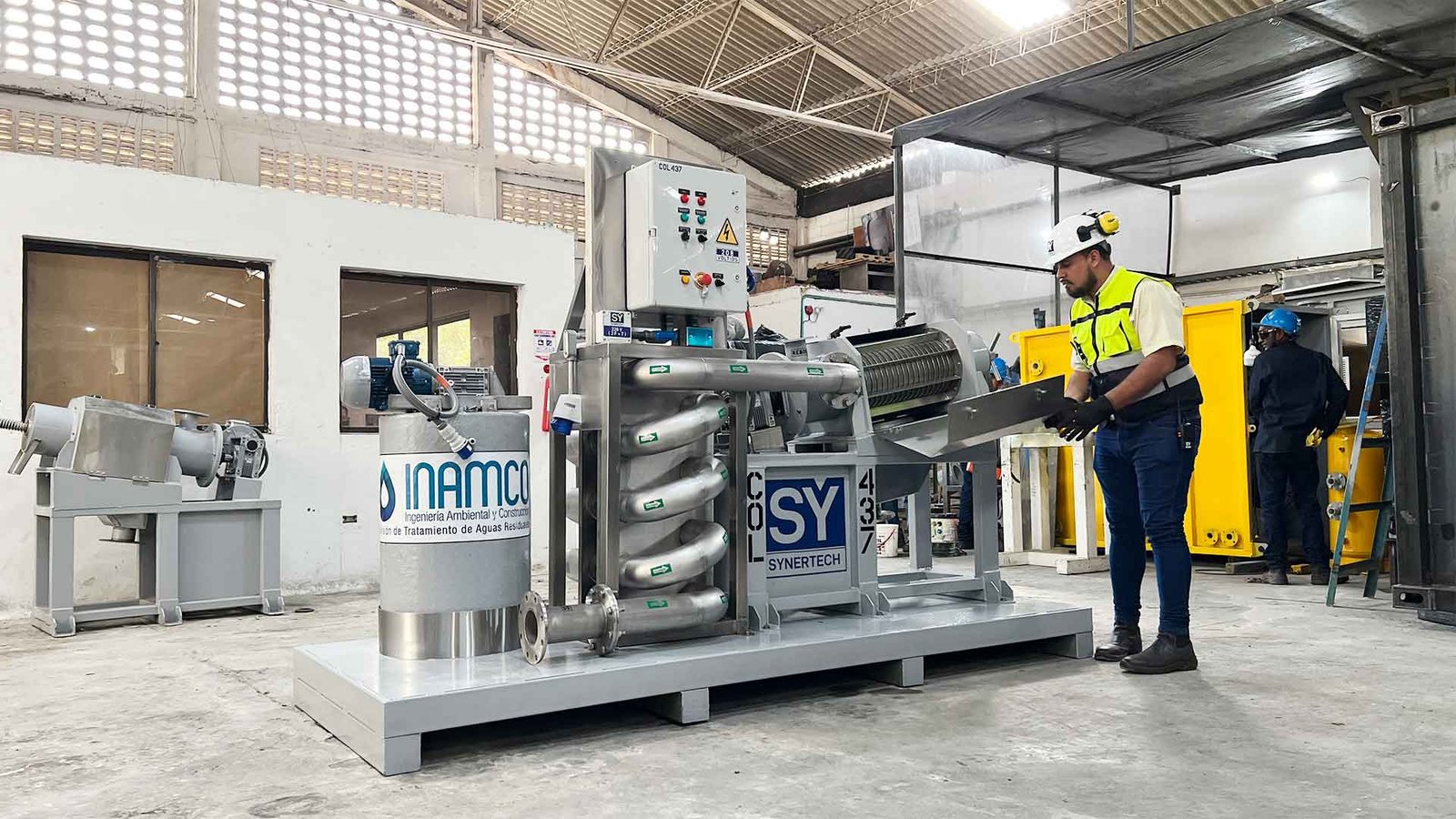 Técnico inspeccionando una planta de tratamiento de aguas residuales modular de color verde marca BIOMAX SYNERTECH, que incluye módulos de pretratamiento, decantación, DAF y oxidación avanzada. La planta está ubicada en un entorno industrial con techo alto y tuberías visibles. - ver más información